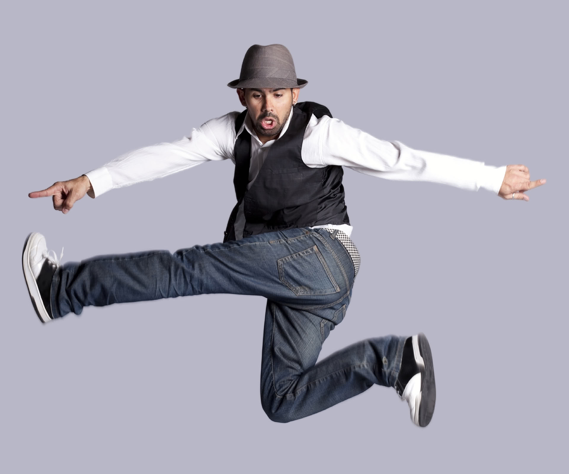 Een jazz danser springt in de lucht met uitgestrekte armen en benen, draagt een hoed, wit overhemd, zwart vest, spijkerbroek en jazz schoenen.